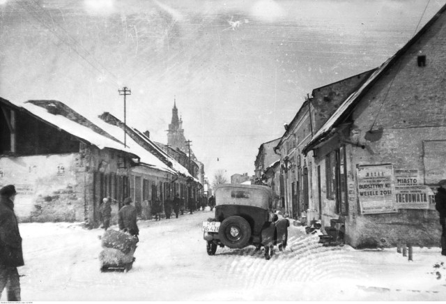 Fragment miasta. Na ulicy widoczni przechodnie i stojący samochód osobowy