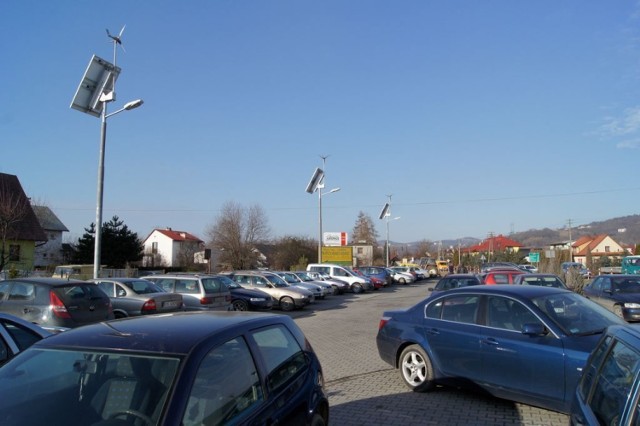 Trzy nowoczesne lampy zasilane hybrydowo (słońce i wiatr) oświetlają nocą parking koło urzędu gminy w Chełmcu.