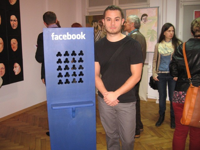 Na honorowym miejscu stoi niebieski konfesjonał z napisem Facebook autorstwa Łukasza Fałdrowicza. Artysta stoi obok pracy.