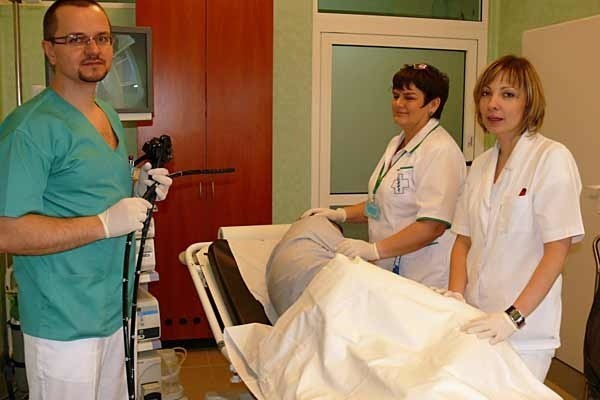 Dr Tomasz Kiszka z pielęgniarkami przy pacjencie przygotowanym do badania wideokolonoskopem, z którego obraz przekazywany jest na monitor.