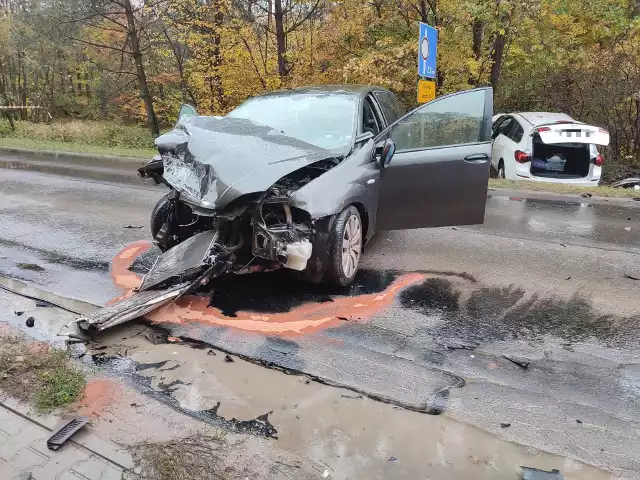 Samochody po kolizji. Przód czarnego auta zniszczony. Wgnieciona karoseria, rozbita szyba, różne elementy na drodze