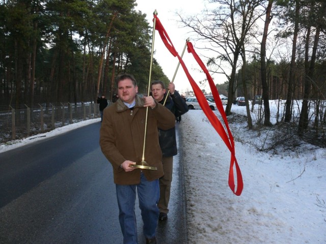 Mężczyźni niosą wstęgę, która za chwilę zostanie ustawiona w poprzek powiatowej drogi bojanowskiej i przecięta na znak zakończenia kolejnej inwestycji.