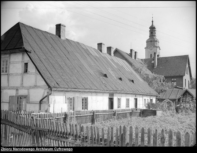 Widok na budynek konstrukcji ryglowej, w tle kościół katolicki pw. Podwyższenia Krzyża Świętego.