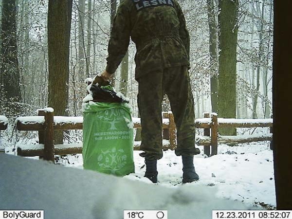 Widok z kamery umieszczonej w lesie przez czaplineckich leśników. Strażnik leśny sprawdza działanie urządzenia w miejscu, w którym wcześniej były już podrzucane śmieci. Na obrazie zarejestrowanym przez kamerę widoczna jest data i godzina nagrania.