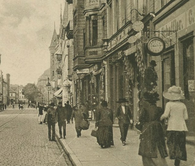 Fotograf uwiecznił zegarmistrza, bądź któregoś z jego pracowników w chwili, gdy ten majstrował coś przy znajdującym się nad witryną zegarze.