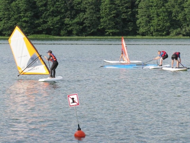 Nauka windsurfingu na jeziorze Głębokie. Młodych adeptów tego sportu asekurują łodzie motorowe. Są poza strefą dla kąpiących się. 