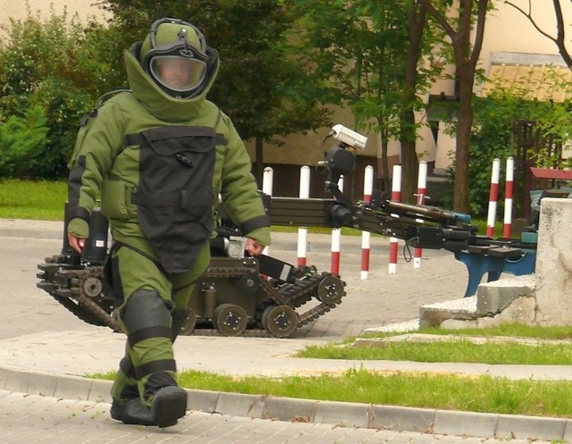 Zdalnie sterowany robot pirotechniczny i policjant z oddziału antyterrorystycznego w skafandrze przeciwodłamkowym pracowali przy niebezpiecznie wyglądającej skrzynce.