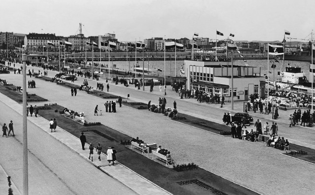 " Gdynia – Aleja Zjednoczenia (ob. Al. Jana Pawła II)Opis Obiektu:Fotografia czarno-biała przedstawia Aleję Zjednoczenia na molo Południowym. Nazwana tak została na cześć zjednoczenia ziem polskich po I wojnie światowej (obecnie al. Jana Pawła II). Z prawej strony, na nabrzeżu Pomorskim, budowana w latach 1937-38 przystań pasażerska Żeglugi Polskiej. W głębi gmachy przy ul. Waszyngtona, m.in. biurowiec Żeglugi Polskiej (obecnie siedziba Dowództwa Floty) oraz Instytut Meteorologii i Gospodarki Wodnej z ażurową wieżą. Na zdjęciu ujęci zostali spacerowicze. Na masztach, wzdłuż Alei, powiewają flagi Żeglugi Polskiej i narodowe. "