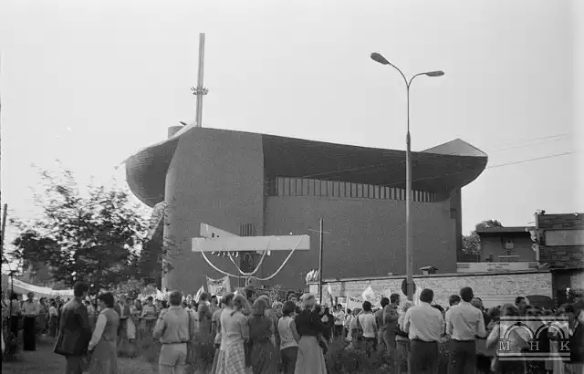 Na ulicy przed kościołem stoją ludzie z transparentami i flagami, oczekujący na przejazd Jana Pawła II do kościoła w Mistrzejowicach. - 1983 r.