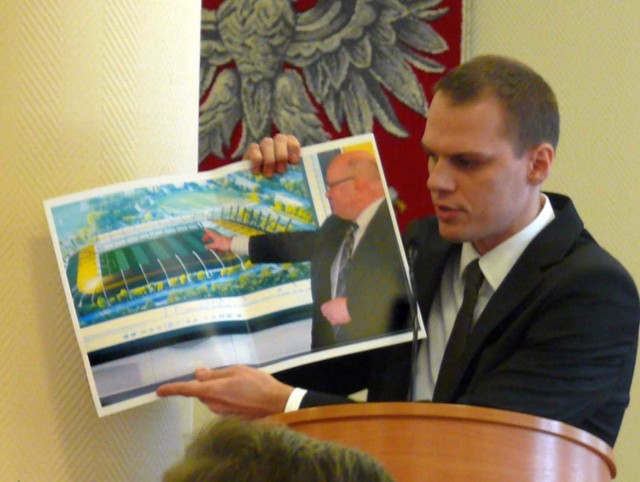 Rafał Weber pokazuje zdjęcie prezydenta obiecującego szybką budowę stadionu piłkarskiego.