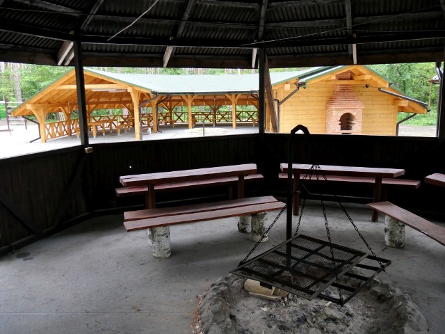 Na pierwszym planie palenisko z grillem, w tle zadaszona wiata ze stołami i ławami z drewna