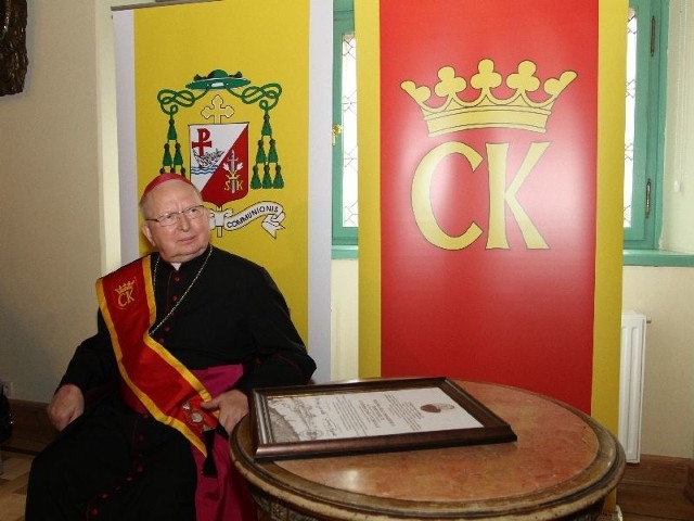 Biskup kielecki, Kazimierz Ryczan już jako Honorowy Obywatel Kielc na tle herbu biskupiego i herbu Kielc, z dokumentem potwierdzającym tytuł.