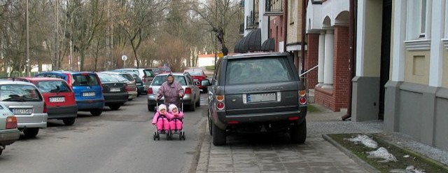 Range rover stoi na chodniku, a kobieta z dwójką dzieci w wózku idzie ulicą.