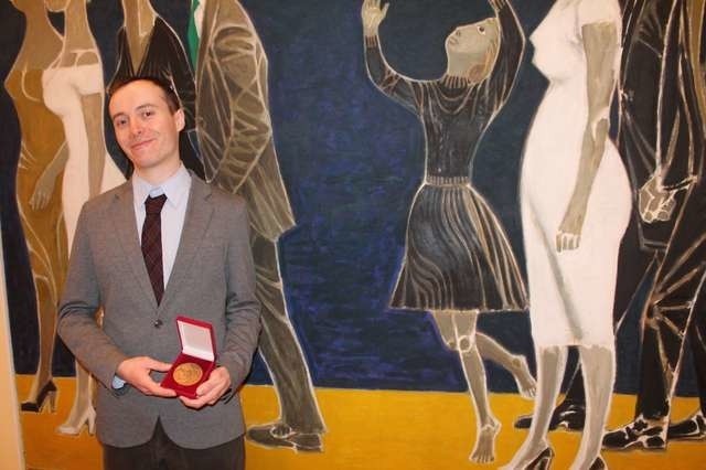 Adam Żuławski z medalem przyznanym przez rektora UMK jego matce, na tle ojcowego obrazu "Ludzie nad morzem", wyeksponowanego w Muzeum Uniwersyteckim
