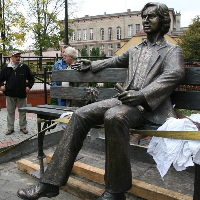 Pomnikowy piosenkarz siedzi na pokrytej jesiennymi liśćmi ławeczce, gestem ręki zapraszając do siebie przechodniów.