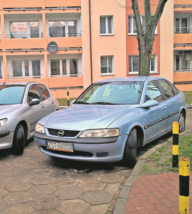 Peugeot i opel zaparkowane na osiedlowej drodze. Z prawej strony przejazd zagradzają słupki, z lewej stoją pojemniki na śmieci