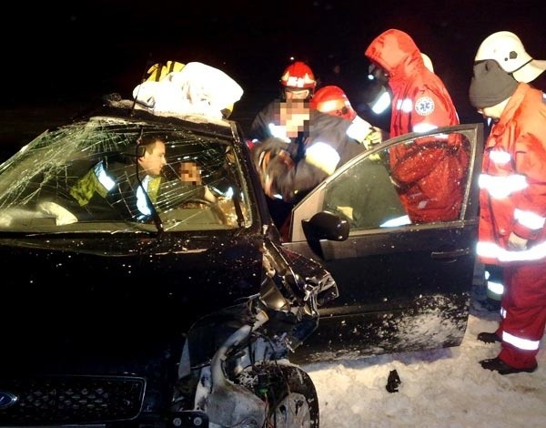 Strażacy wyciągają 31-latkę z rozbitego auta. Za nimi czeka ekipa pogotowia, aby odwieźć poszkodowaną do szpitala.