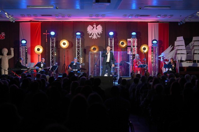 Koncert Bardów Wolności w historycznej sali BHP Stoczni Gdańskiej. Artyści na scenie, liczna publiczność na widowni.