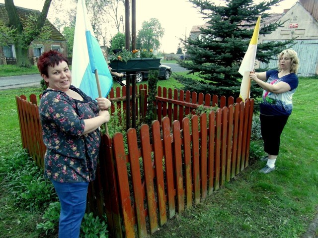 Sołtys Drzeńska Weronika Krasucka razem z Mariolą Białą wieszają flagi przy krzyżu, który stoi we wsi.