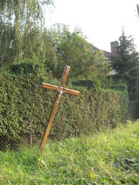 Kilka metrów przed furtką przechyla się drewniany, nagrobny krzyż. Lakier miejscami się łuszczy, pod figurką Jezusa jest ślad po tabliczce z nazwiskiem zmarłego.