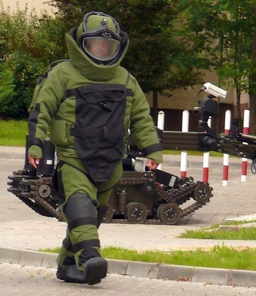 Zdalnie sterowany robot pirotechniczny i policjant z oddziału antyterrorystycznego w skafandrze przeciwodłamkowym pracowali przy niebezpiecznie wyglądającej skrzynce.