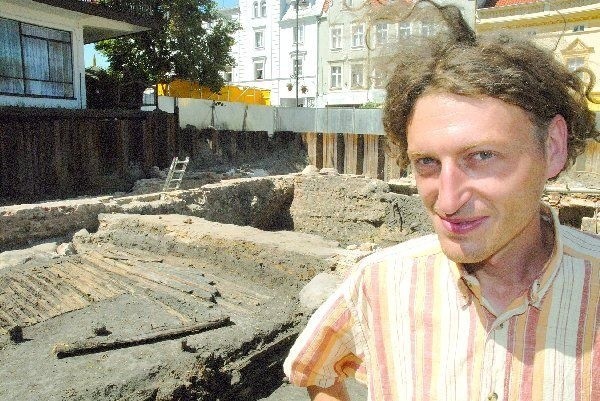 Archeolog Wojciech Siwiak na tle  drewnianych pozostałości zabudowań  gospodarczych z XIV-XV w.
