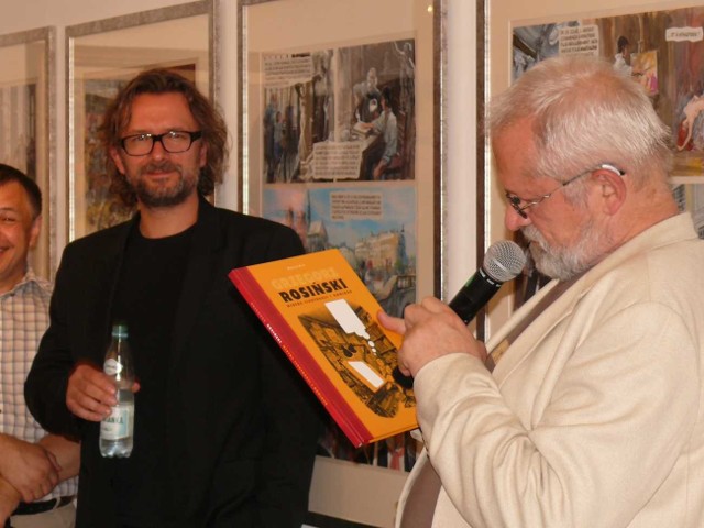 Grzegorz Rosiński na otwarciu wystawy, w ręce trzyma katalog. Oboj jego syn Piotr, komisarz wystawy.