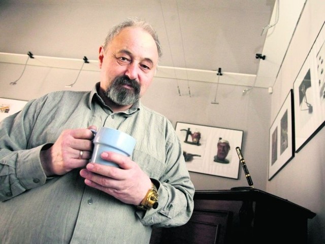 Marek Mazur w swojej pracowni z kubkiem herbaty i złoconym naręcznym aparatem fotograficznym. Na komodzie wieczne pióro z kamerą filmową