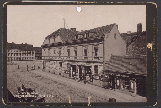 Zdjęcie z 1897 r., wydane przez Albina Johanssmanna, gnieźnieńskiego wydawcę i księgarza. Wylot obecnej ul. Chrobrego (wówczas – Friedrichstrasse) na Rynek w Gnieźnie. Narożny, istniejący do dziś budynek, mieścił antykwariat i księgarnię wydawcy tego zdjęcia, po jego śmierci w 1898 r. prowadzone do 1905 r. przez wdowę po nim, Paulę. Parterowy dom z zakładem fryzjerskim zniknął na przełomie wieków.