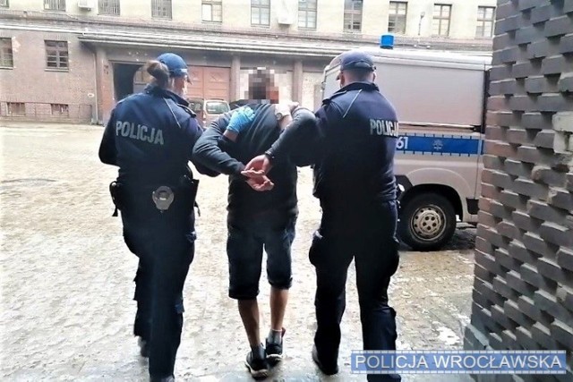 Zatrzymany mężczyzna z kajdankami na rękach prowadzony przez umundurowanych policjantów w kierunku oznakowanego radiowozu.