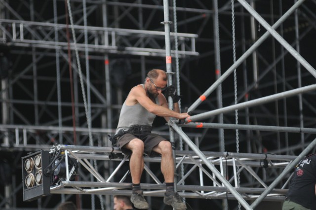 Prace na terenie PolAndRock Festiwalu idą pełną parą. Miejsce, które jeszcze do niedawna było zwykłą, trawiastą łąką, odwiedziliśmy we wtorek, 24 lipca. Ale tu się dzieje! Wszędzie pracują ludzie, jeździ sprzęt, dowożone są nowe elementy festiwalowej infrastruktury, budowane są nowe namioty, rozstawiane kontenery. Ale nad całością dominuje Duża Scena. Zobaczcie, jak idzie jej budowa.



To właśnie pod Dużą Sceną składowane są kontenery, które później rozwożone są we wszystkie strony woodstockowego pola. Stoi tu też część ogrodzeń, którymi będą odgrodzone najważniejsze miejsce na polu i sam teren festiwalu. Przy Dużej Scenie nie zobaczycie jednak w tym roku barierek. I jest to informacja, która cieszy chyba wszystkich uczestników PolAndRock Festiwalu. 

Przy scenie spotkaliśmy członków Pokojowego Patrolu. Jak zwykle to właśnie oni, czyli kwaterka, zajmują się m. in. obijaniem deskami frontu sceny. Prace idą dość sprawnie, ale utrudnia je lejący się z nieba żar. Bywa, że jest tu nawet 40 stopni! Obecnie w Kostrzynie jest 12 członków Pokojowego Patrolu. To ludzie, którzy pomagają w przygotowaniu wszystkiego na przyjazd kolejnych "patrolowców" i samych uczestników imprezy. 

