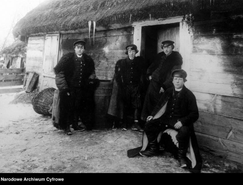 Grupa mężczyzn w strojach ludowych przed chatą.