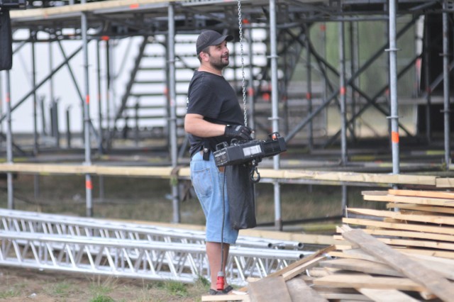 Prace na terenie PolAndRock Festiwalu idą pełną parą. Miejsce, które jeszcze do niedawna było zwykłą, trawiastą łąką, odwiedziliśmy we wtorek, 24 lipca. Ale tu się dzieje! Wszędzie pracują ludzie, jeździ sprzęt, dowożone są nowe elementy festiwalowej infrastruktury, budowane są nowe namioty, rozstawiane kontenery. Ale nad całością dominuje Duża Scena. Zobaczcie, jak idzie jej budowa.



To właśnie pod Dużą Sceną składowane są kontenery, które później rozwożone są we wszystkie strony woodstockowego pola. Stoi tu też część ogrodzeń, którymi będą odgrodzone najważniejsze miejsce na polu i sam teren festiwalu. Przy Dużej Scenie nie zobaczycie jednak w tym roku barierek. I jest to informacja, która cieszy chyba wszystkich uczestników PolAndRock Festiwalu. 

Przy scenie spotkaliśmy członków Pokojowego Patrolu. Jak zwykle to właśnie oni, czyli kwaterka, zajmują się m. in. obijaniem deskami frontu sceny. Prace idą dość sprawnie, ale utrudnia je lejący się z nieba żar. Bywa, że jest tu nawet 40 stopni! Obecnie w Kostrzynie jest 12 członków Pokojowego Patrolu. To ludzie, którzy pomagają w przygotowaniu wszystkiego na przyjazd kolejnych "patrolowców" i samych uczestników imprezy. 

