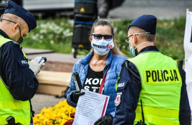 Trzech policjantów próbuje legitymować niewidomą Dorotę Halaburdę przed pomnikiem króla w trzeciomajowe święto.