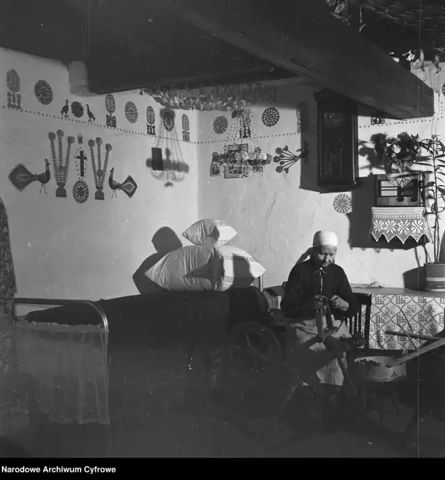Wnętrze chaty kurpiowskiej, tzw. biała izba. Widoczna Marianna Konopka pracująca przy kołowrotku. Na ścianach wiszą ludowe wycinanki z papieru (tzw. leluje), lata 1960-1970