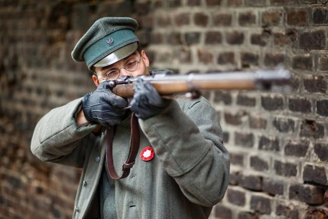 Powstaniec Wielkopolski gotowy do strzałuMundur Powstańca Wielkopolskiego. Spodnie, wzór 1910, pochodziły z typowego umundurowania armii pruskiej z początku I Wojny Światowej. Charakterystyczne owijacze na spodniach były stosowane przed pojawieniem się wysokich butów. Było to rozwiązanie ekonomiczne, które się względnie sprawdzało. W okopach ziemia nie wpadała do trzewików, ale owijacze przemarzały. Używano niemieckich kurtek podoficerskich i oficerskich wzór 1915. Czasami były z nich odpruwane guziki. Pas z klamrą też były niemieckie – wszystko wykorzystano. Zamiast hełmów zakładano feldmyce. Karabiny w większości były niemieckie. Zdarzały się również francuskie – przejęte na froncie – problemem była amunicja do nich.Płaszcz służył do wszystkiego. Do chodzenia, jako koc do spania – był z bardzo wytrzymałego materiały. Kolor munduru nie był przypadkowy, bo stosowano kamuflaż.