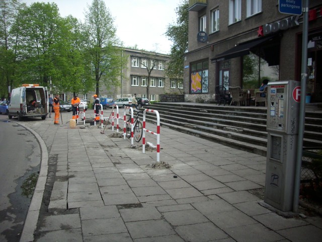 Ekipa drogowa montuje bariery-stojaki rowerowe na szerokim chodniku koło kawiarni.