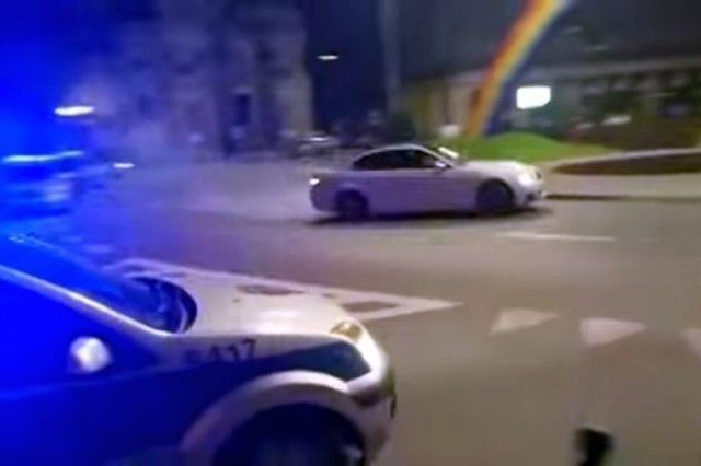 BMW Froga driftuje na Placu Zbawiciela w Warszawie. W tle widoczna Tęcza. Na pierwszym planie próbujący interweniować radiowóz.
