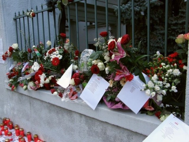 Kwiaty zatknięte za ogrodzenie ambasady, a wśr&oacute;d nich kartki z listami ofiar katastrofy, podzielonymi według pełnionej służby.