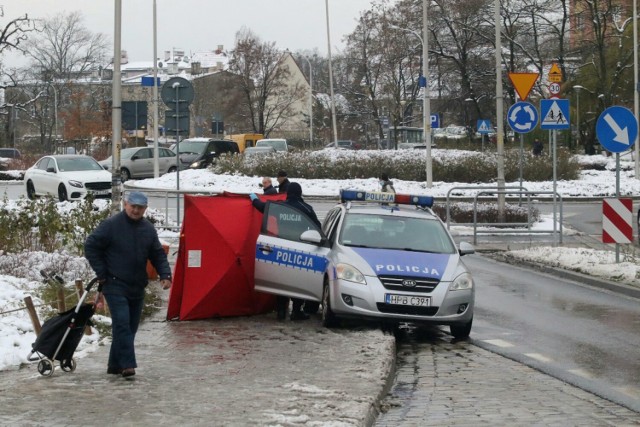 Policja zabezpiecza zwłoki rowerzysty leżące na skraju chodnika i jezdni, w pobliżu przystanku autobusowego. 