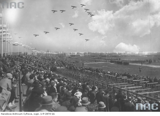 3 maja 1936 r. Rewia wojskowa na Polu Mokotowskim. Widok ogólny trybun dla publiczności oraz placu wypełnionego oddziałami wojskowymi biorącymi udział w uroczystości. Nad terenem uroczystości przelatuje eskadra samolotów wojskowych.