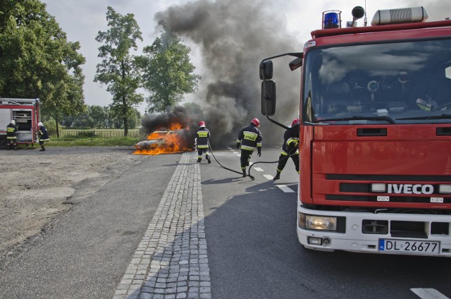 Fotografia przedstawiająca płonący samochód oraz interwencję Straży Pożarnej dnia 04.08 w Kochlicach.